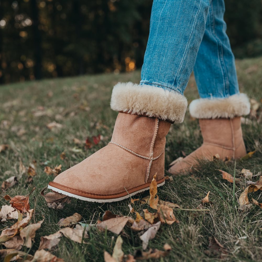 A photo of Zeezoo Dingo made with suede, sheepskin, and rubber soles. The boots are brown in color with tan soles and an ugg style look. Both boots are shown diagonally from the front left on a woman’s feet, with a view of her knees down. The cuff of the boots is folded down to show the sheepskin interior. The woman is wearing blue jeans tucked into the boots and is walking in grass with her left foot in front of her right. #color_chestnut