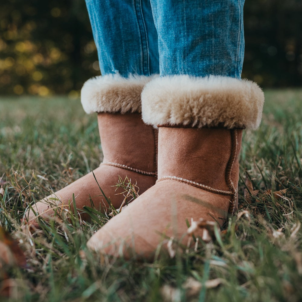 A photo of Zeezoo Dingo made with suede, sheepskin, and rubber soles. The boots are brown in color with tan soles and an ugg style look. Both boots are shown diagonally from the front left on a woman’s feet, with a view of her knees down. The cuff of the boots is folded down to show the sheepskin interior. The woman is wearing blue jeans tucked into the boots and is standing in grass. #color_chestnut