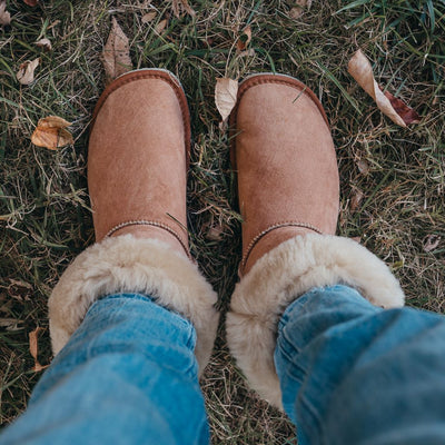 A photo of Zeezoo Dingo made with suede, sheepskin, and rubber soles. The boots are brown in color with tan soles and an ugg style look. Both boots are shown diagonally from above on a woman’s feet, with a view of her shins down. The cuff of the boots is folded down to show the sheepskin interior. The woman is wearing blue jeans tucked into the boots and is standing in grass. #color_chestnut