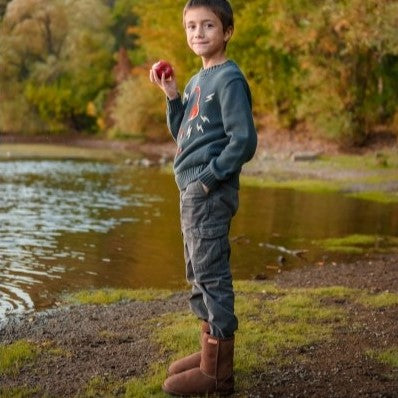 A photo of kids Zeezoo dingo boots made with suede lined with sheepskin and rubber soles. The boots are brown in color and they go around the mid calf. Both boots are shown on a little boy eating an apple standing on a grassy patch near water with green foliage in the background. #color_brown