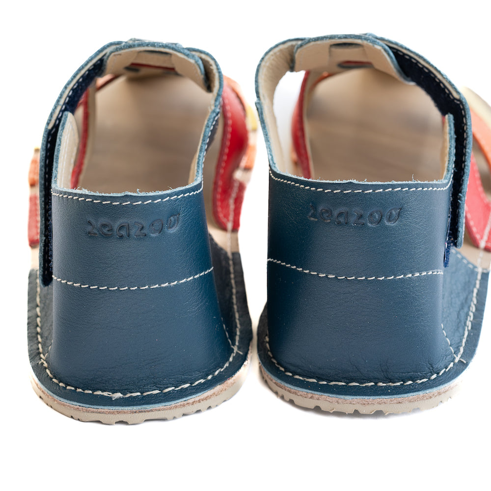 A photo of the Zeazoo Marlin lids leather play sandals. The sandals are navy blue in color and have yellow, orange, and red straps weaved across the top of the shoe, and a velcro closure around the ankle. Both shoes are shown from behind on a white background, displaying the Zeazoo logo on the heels. #color_navy-yellow-orange-red