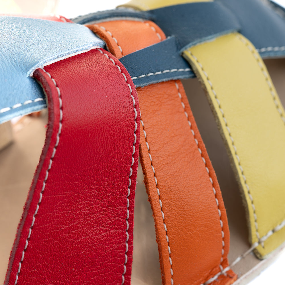 A photo of the Zeazoo Marlin lids leather play sandals. The sandals are navy blue in color and have yellow, orange, and red straps weaved across the top of the shoe, and a velcro closure around the ankle. A close up of the colored straps is shown from the right side on a white background. #color_navy-yellow-orange-red