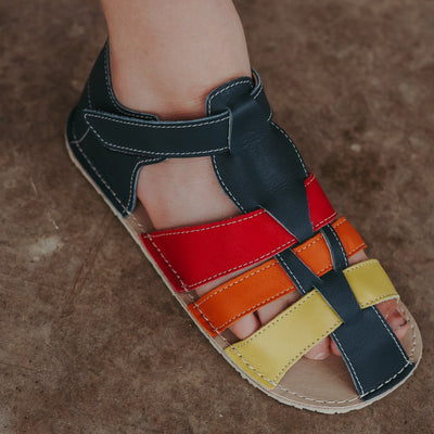 A photo of the Zeazoo Marlin lids leather play sandals. The sandals are navy blue in color and have yellow, orange, and red straps weaved across the top of the shoe, and a velcro closure around the ankle. Right shoe is shown facing diagonally right on a little kid standing on cement. #color_navy-yellow-orange-red