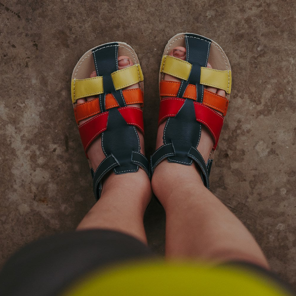 A photo of the Zeazoo Marlin lids leather play sandals. The sandals are navy blue in color and have yellow, orange, and red straps weaved across the top of the shoe, and a velcro closure around the ankle. Both shoes are shown from above on a little kid standing on cement. #color_navy-yellow-orange-red