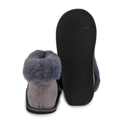 Grey colored Zeezoo Shaun hand made suede, sheepskin, and tan rubber slippers. The slippers are composed of 4 panels sewn together with seams on the sides, back, and top. Fur lining folds over at the ankle. Left slipper is shown from the back next to the right slipper sole facing up against a white background. #color_grey