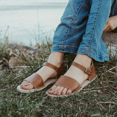 A photo of Brown Zeezoo Siren leather Sandals. The sandals have a front foot strap, a leather ankle strap, and a heel cup. Both sandals are shown diagonally left here. Shoes are worn by a young woman sitting on a rock with her feet in the grass and a small body of water in the background. Woman is wearing medium-wash flare jeans. #color_brown