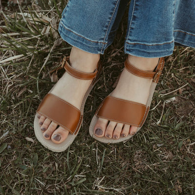 A photo of Brown Zeezoo Siren leather Sandals. The sandals have a front foot strap, a leather ankle strap, and a heel cup. Both sandals are shown from above here facing diagonally left. Shoes are worn by a young woman sitting in the grass wearing medium-wash flare jeans. #color_brown