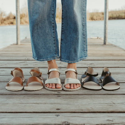 A photo of Brown Zeezoo Siren leather Sandals. The sandals have a front foot strap, a leather ankle strap, and a heel cup. Both sandals are shown from above here facing diagonally left. Gold sandals are shown here worn by a woman standing on a narrow dock wearing wide-legged, medium-wash, cropped jeans. Brown sandals are to the left of the woman and black sandals are to the right with a small body of water and a tree line in the background. #color_brown