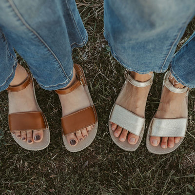 A photo of Gold Zeezoo Siren leather Sandals. The sandals have a front foot strap, a leather ankle strap, a heel cup, cream soles, and a cream footbed. Both sandals are shown here from above on a woman standing in grass wearing wide-legged, medium-wash, cropped jeans. A young woman is standing to the left of her wearing the brown siren model and similar jeans. #color_gold