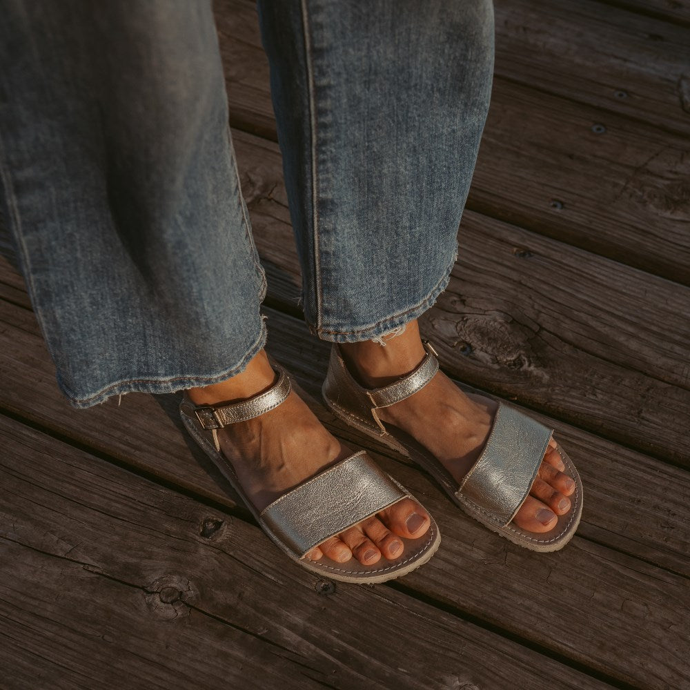 A photo of Gold Zeezoo Siren leather Sandals. The sandals have a front foot strap, a leather ankle strap, a heel cup, cream soles, and a cream footbed. Both sandals are shown here diagonally right on a woman standing on a wooden dock wearing wide-legged, medium-wash, cropped jeans. #color_gold