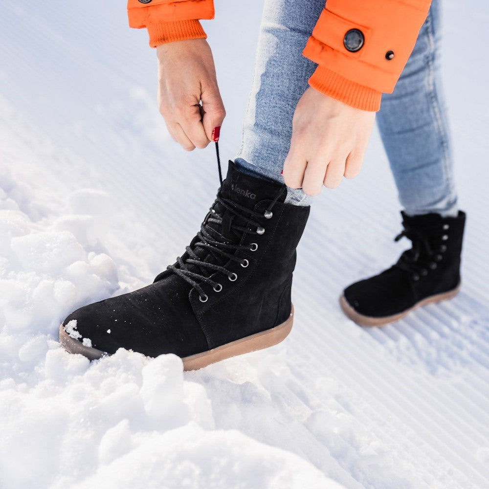 A photo of Be Lenka Winter Neo boots made with leather and rubber soles. The boots are black in color and a lace up style with wool inside. Left boot is shown atop a snow pile with a woman tying the boot. Woman is wearing lightwash skinny jeans and an orange winter coat standing in snow. #color_matte-black-nubuck