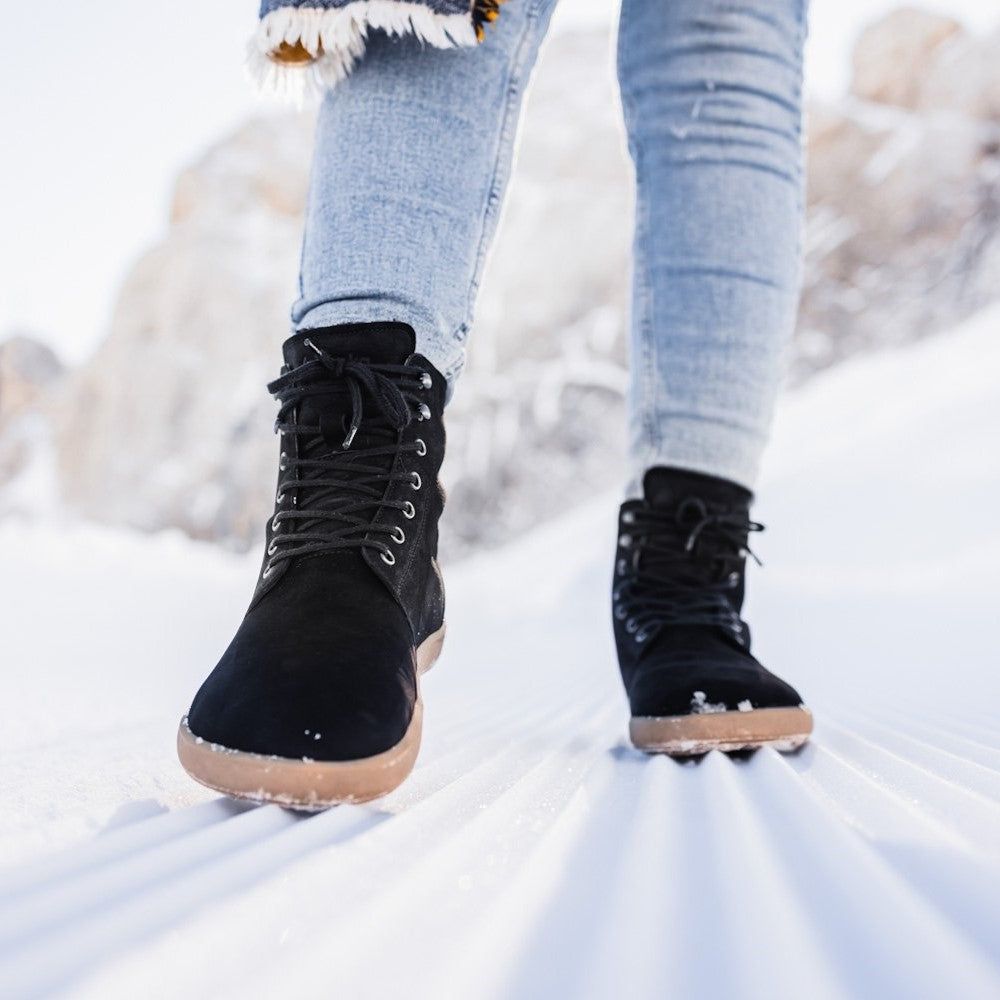 A photo of Be Lenka Winter Neo boots made with leather and rubber soles. The boots are black in color and a lace up style with wool inside. Both boots are shown from the front on a woman wearing lightwash skinny jeans standing in snow. #color_matte-black-nubuck
