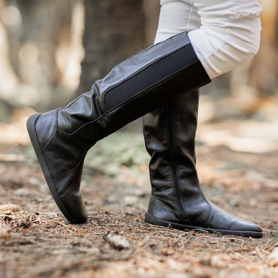 A photo of Belenka Sierra made from smooth leather, fleece, and rubber soles. The boots are black in color with a tall riding boot elastic paneled shaft lined with fleece. A woman is shown standing in the woods wearing tan pants tucked into the Sierra boots, she has her right leg slightly bent. #color_black