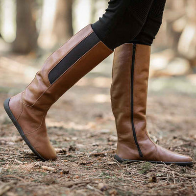 A photo of Belenka Sierra made from smooth leather, fleece, and rubber soles. The boots are brown in color with a tall riding boot elastic paneled shaft lined with fleece. A woman is shown standing in the woods wearing tan pants tucked into the Sierra boots, she has her right leg slightly bent. #color_brown