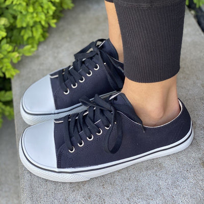 A photo of Bohempia Herlik canvas sneakers made from canvas and rubber soles. The sneakers are a black color with a white toe cap and a black outline around the white rubber soles. Both sneakers are shown together from the left side on a woman’s feet with a view of her shins down. The woman is wearing   black jogger pants and is standing on a cement step. #color_black-white