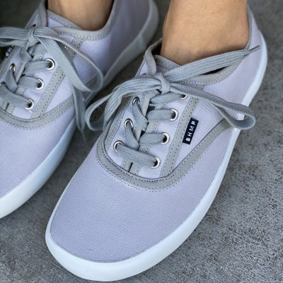 A photo of Bohempia Kolda Plimsole sneakers made from canvas and rubber soles. The sneakers are grey in color with trim detailing and a small tag on the top by the laces. A woman is shown standing wearing the shoes an up close shot is shown of the right shoe and the left shoe is part cut out of the picture. #color_grey