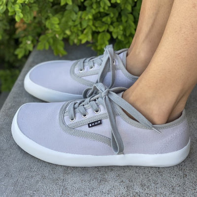 A photo of Bohempia Kolda Plimsole sneakers made from canvas and rubber soles. The sneakers are grey in color with trim detailing and a small tag on the top by the laces. A woman is shown from mid leg down wearing the velik sneakers standing sideways to the left on concrete steps with greenery in the background. #color_grey