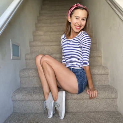 A photo of Bohempia Kolda Plimsole sneakers made from canvas and rubber soles. The sneakers are grey in color with trim detailing and a small tag on the top by the laces. A brown haired woman is shown smiling sitting on brown carpeted steps wearing a red hair wrap, blue and white striped shirt, jean shorts, and the Kolda sneakers. #color_grey