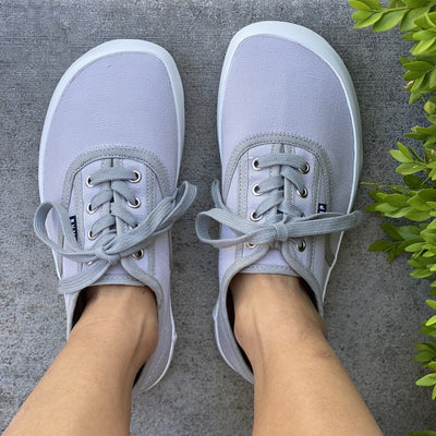 A photo of Bohempia Kolda Plimsole sneakers made from canvas and rubber soles. The sneakers are grey in color with trim detailing and a small tag on the top by the laces. A woman is shown above from the mid leg down wearing the herlik sneakers standing on concrete steps. #color_grey