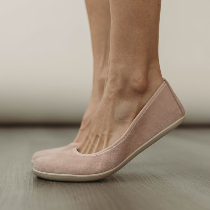 A photo of Groundies Lily Soft flats with a leather upper and cream rubber true sense soles. The flats are a nubuck in a light pink color, the trim around the tops of the flats is a light pink color that is lighter than the rest of the flats. The interior of the flats is a light beige color. A woman is shown from mid leg down wearing the Lily flats and standing with her heels lifted on a grey wood floor with a cream wall in the background. #color_light-pink