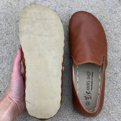 A photo of Yemeni Leather Loafers Designed by Anya with a leather upper and soles. The loafers are a brown color with tan leather soles they have small slits on each side with elastic on the top of the foot. Both shoes are shown on a concrete background with a woman’s hand holding the left shoe upside down to show the leather sole. #color_brown