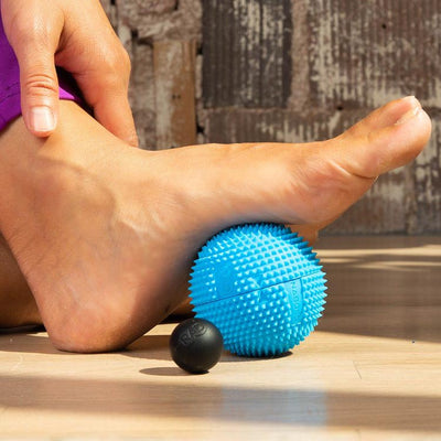 A photo of Naboso neuro ball that can be split in half and contains a smaller RAD ball inside. The blue is blue in color and textured and can be used on the feet. A woman is shown sitting on a wood floor from the ankle down wearing purple leggings and her hand resting on her ankle while rolling her foot on the neuro ball. 