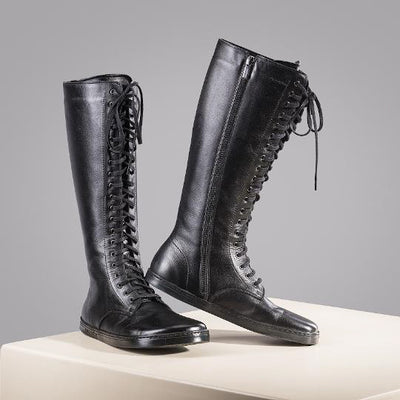 A photo of Peerko Empire boots made from leather and rubber soles. The boots are black in color, they are a tall riding boot style with laces all the way up and a side zipper. Both boots are shown sitting on a white box angled slightly to the right, the left boot has its heel leaned on the right boot against a gray background. #color_black