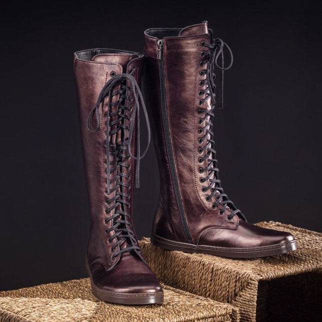 A photo of Peerko Empire boots made from leather and rubber soles. The boots are chestnut metallic in color, they are a tall riding boot style with laces all the way up and a side zipper. Both boots are shown sitting on two sisal boxes angled slightly to the right, the left boot has its heel leaned on the right boot against a black background. #color_chestnut
