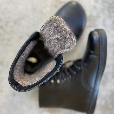A photo of Peerko Frost combat boots made with smooth leather, wool, and rubber soles. The boots are black in color, wool lined, with silver speed hooks at the top. Both boots are shown from above the left foot is lying on it’s side and the right boot is sitting upright with the tongue folded down to show the wool lined inside against a concrete background. #color_black