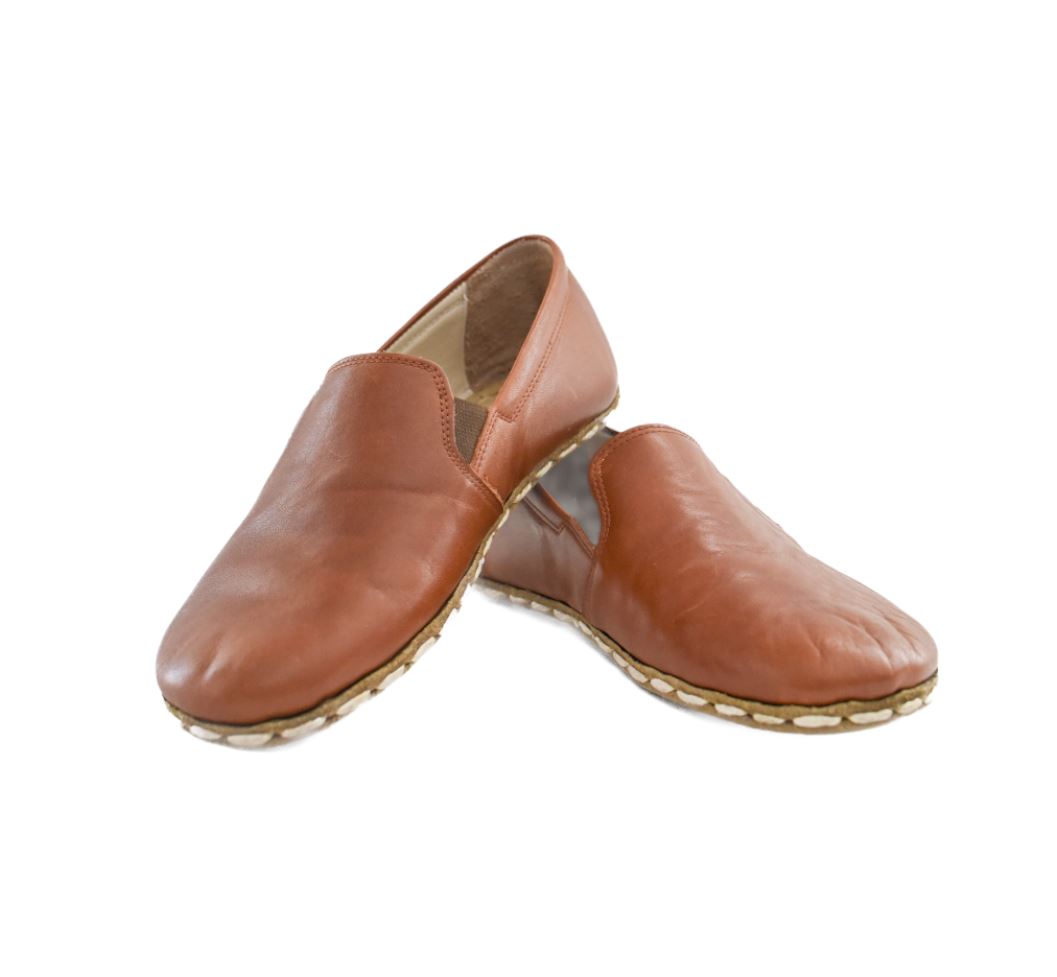A photo of Yemeni Leather Loafers Designed by Anya with a leather upper and soles. The loafers are a brown color with tan leather soles they have small slits on each side with elastic on the top of the foot. Both loafers are shown facing opposite directions with the heel of the right shoe is leaning on the left shoe against a white background. #color_brown