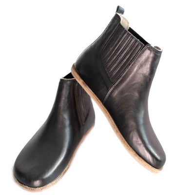 A photo of designed by Anya Lila boots made from leather and rubber soles. The boots are black in color, they are a Chelsea boot style with a lined detailed panel on the sides. Both boots are shown the right boot is shown from the front angled slightly to the left, the heel of the is resting on the left boot and facing downward against a white background. #color_black-smooth-leather