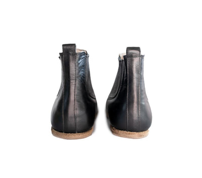 A photo of designed by Anya Lila boots made from leather and rubber soles. The boots are black in color, they are a Chelsea boot style with a lined detailed panel on the sides. Both both are shown beside each other from the back heel against a white background. #color_black-smooth-leather