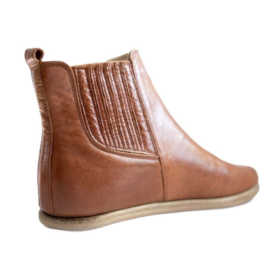 A photo of designed by Anya Lila boots made from leather and rubber soles. The boots are black in color, they are a Chelsea boot style with a lined detailed panel on the sides. One boot is shown angled from the back right against a white background. #color_brown