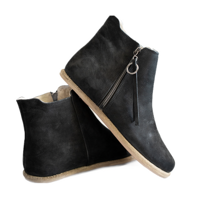 A photo of designed by Anya Rosa boots made from nubuck leather and rubber soles. The boots are black in color, they are a Chelsea boot style with a zipper on the sides. Both boots are shown the left boot is shown from the back angled slightly to the left, the heel of the is resting on the right boot and facing downward against a white background. #color_black-nubuck