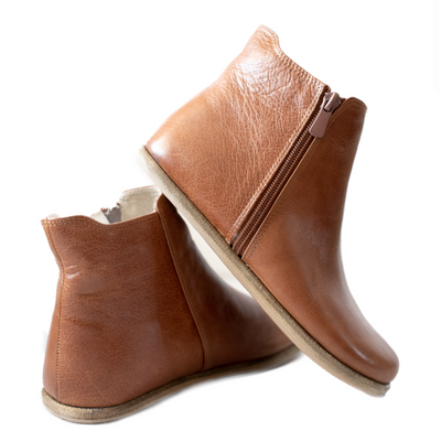 A photo of designed by Anya Rosa boots made from leather and rubber soles. The boots are brown in color, they are a Chelsea boot style with a zipper on the sides. Both boots are shown the right boot is shown from the back, the heel of the is resting on the left boot and facing downward against a white background. #color_brown