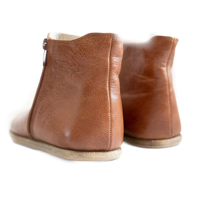 A photo of designed by Anya Rosa boots made from leather and rubber soles. The boots are brown in color, they are a Chelsea boot style with a zipper on the sides. Both boots are shown beside each other up close from the back heel against a white background. #color_brown