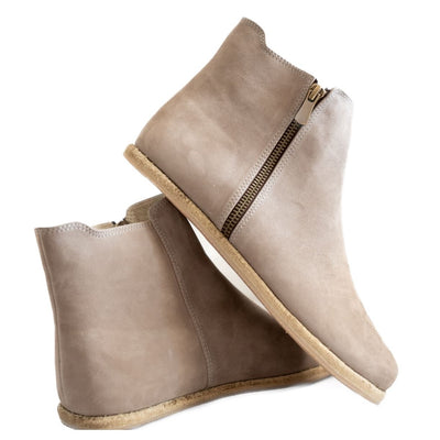 A photo of designed by Anya Rosa boots made from nubuck leather and rubber soles. The boots are taupe in color, they are a Chelsea boot style with a zipper on the sides. Both boots are shown the left boot is shown from the back angled slightly to the left, the heel of the is resting on the right boot and facing downward against a white background. #color_taupe-nubuck