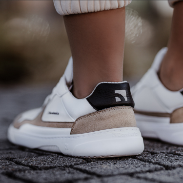 A photo of Barebarics Zing sneakers made with a leather upper and a rubber sole. The sneakers are a white and beige color with perforated spots on the top of the toe box and barebarics branding on the tongue and side. They also have black at the top of the heel. A person is shown from the mid leg down standing wearing the shoes with their heels towards us the photo is angled to show the back of the left shoe closer on a concrete background. #color_white-beige
