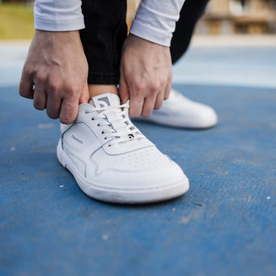 A photo of Barebarics Zing sneakers made with a leather upper and a rubber sole. The sneakers are a white color with perforated spots on the top of the toe box and barebarics branding on the tongue and side. A person is shown standing from the mid-leg down wearing black pants and the sneakers, she is holding both sides of the shoe beside the tongue against a blue background with blurry grass in the distance. #color_white