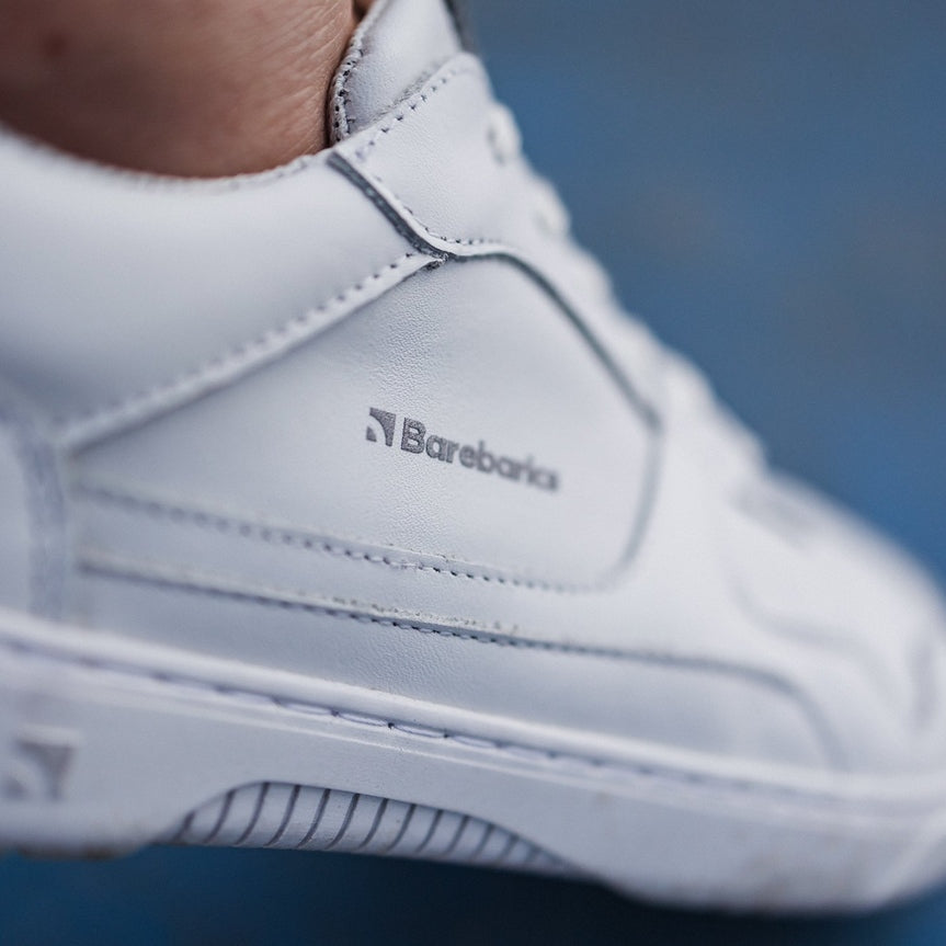 A photo of Barebarics Zing sneakers made with a leather upper and a rubber sole. The sneakers are a white color with perforated spots on the top of the toe box and barebarics branding on the tongue and side. A person is shown wearing the sneaker up close to show the shoes detail and branding against a blue background. #color_white