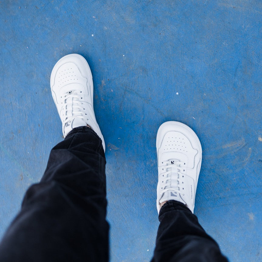 A photo of Barebarics Zing sneakers made with a leather upper and a rubber sole. The sneakers are a white color with perforated spots on the top of the toe box and barebarics branding on the tongue and side. A person is shown standing mid-thigh down wearing black jogging pants and the sneakers against a blue background. #color_white