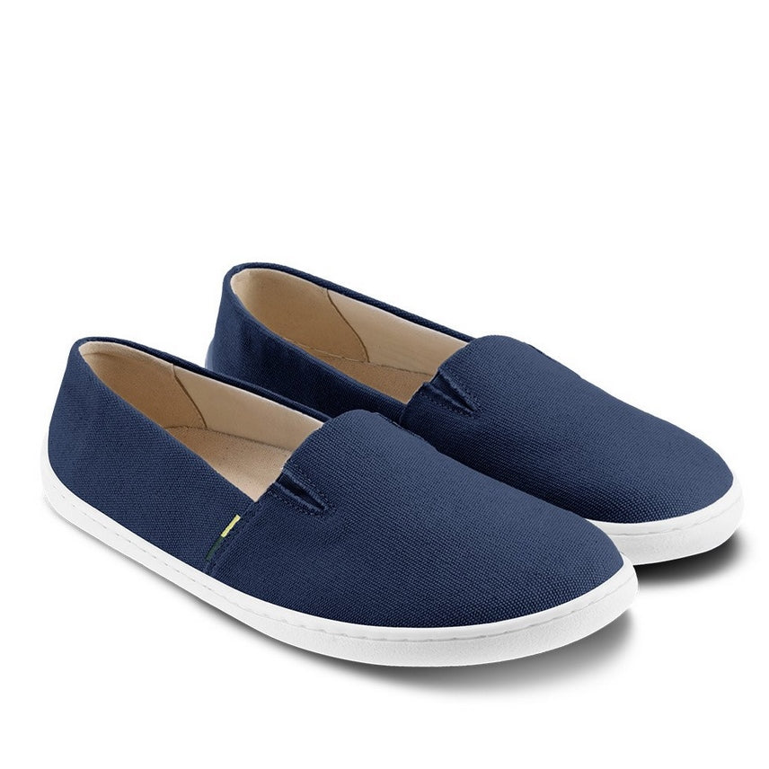A photo of Dark Blue Be Lenka Bali canvas loafers with rubber soles. The loafers have two V-shaped stitching details at the top opening of the shoe. Both shoes are shown with the left shoe facing right and the right shoe heel propped up on the left shoe against a white background. #color_dark-blue