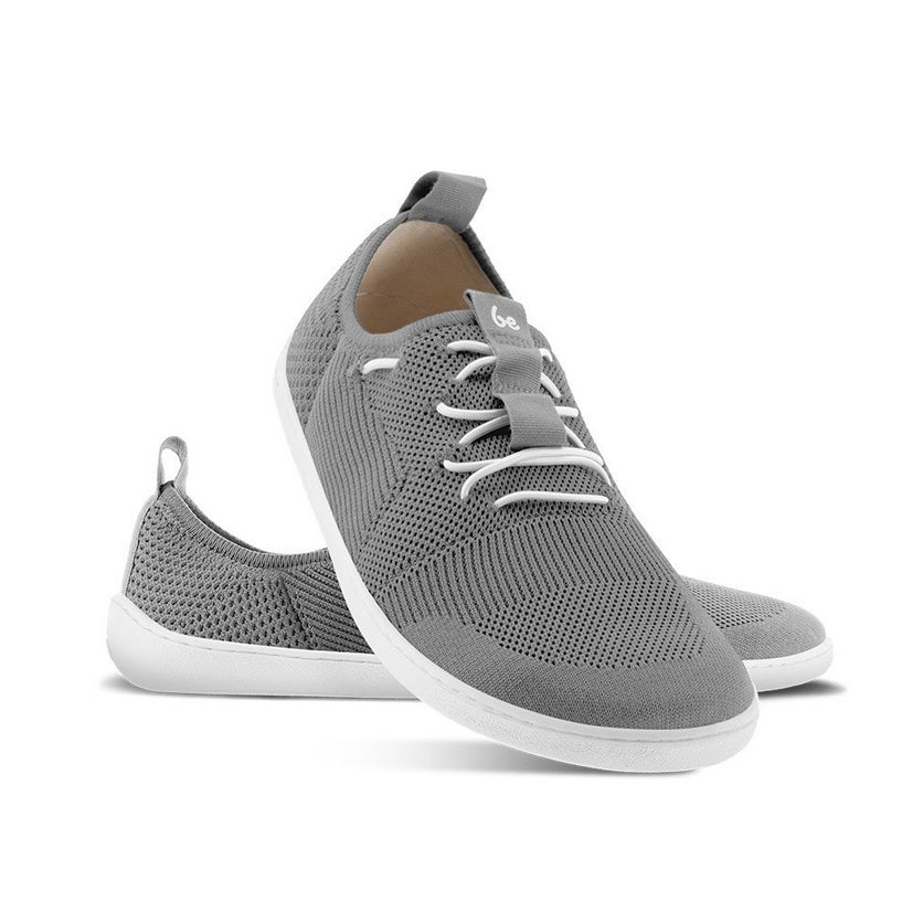 A photo of Grey Be Lenka Elevate Mesh Sneaker shoes with white soles and white elastic laces. Left shoe is shown from the right with the right shoe propped up on the left to show the top of the shoe. Background is white. #color_grey