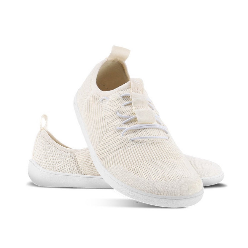 A photo of Ivory White Be Lenka Elevate Mesh Sneaker shoes with white soles and white elastic laces. Left shoe is shown from the right with the right shoe propped up on the left to show the top of the shoe. Background is white. #color_ivory-white