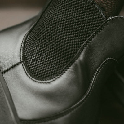 A photo of Belenka Entice Neo boots made from smooth leather and black rubber soles. The boots are black in color with elastic panels on the sides and pull on loops. One boot is shown up close from the side to shown the detail of the elastic. #color_black
