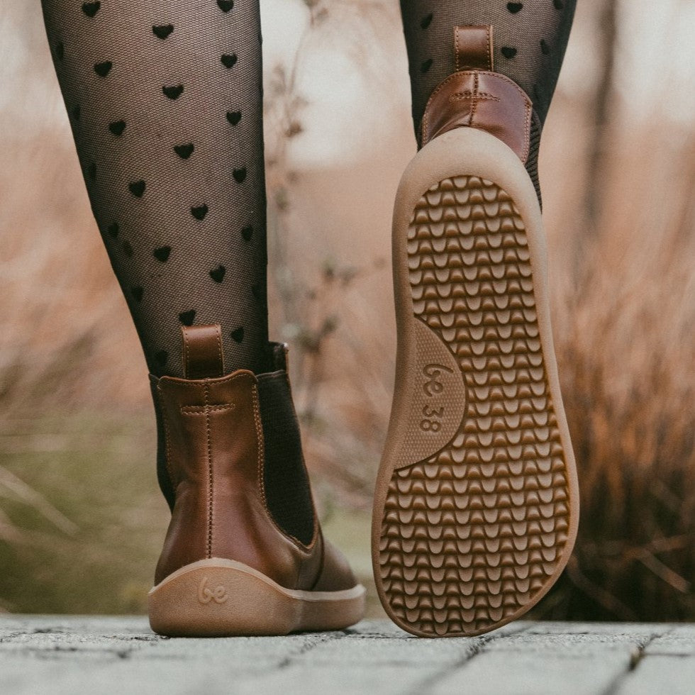A photo of Belenka Entice Neo boots made from smooth leather and tan rubber soles. The boots are brown in color with dark brown elastic panels on the sides and pull on loops. A woman is shown from mid leg down walking down a path of concrete blocks wearing black sheer heart tights and the boots the left shoes sole is showing while she takes a step there is tall tan grasses in the background. #color_dark-brown
