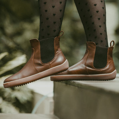 A photo of Belenka Entice Neo boots made from smooth leather and tan rubber soles. The boots are brown in color with dark brown elastic panels on the sides and pull on loops. A woman is shown walking down steps from the mid-leg down wearing black sheer heart tights and the boots there is greenery in the background. #color_dark-brown