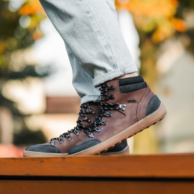 A photo of Be Lenka Ranger boots made with oiled nubuck leather, fleece, and rubber soles. The boots are a hiking boot style and are brown in color with black trim and a fleece lining. A woman is shown from mid leg down wearing rolled jeans and the ranger boots while standing on a wood porch with some blurry trees in the distance. #color_dark-brown