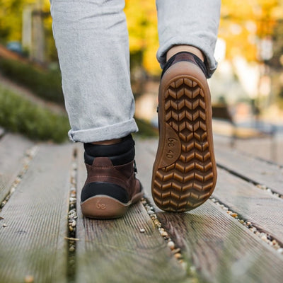 A photo of Be Lenka Ranger boots made with oiled nubuck leather, fleece, and rubber soles. The boots are a hiking boot style and are brown in color with black trim and a fleece lining. A woman is shown walking on a wood porch from mid leg down wearing rolled jeans and the ranger boots with blurry trees in the distance. #color_dark-brown