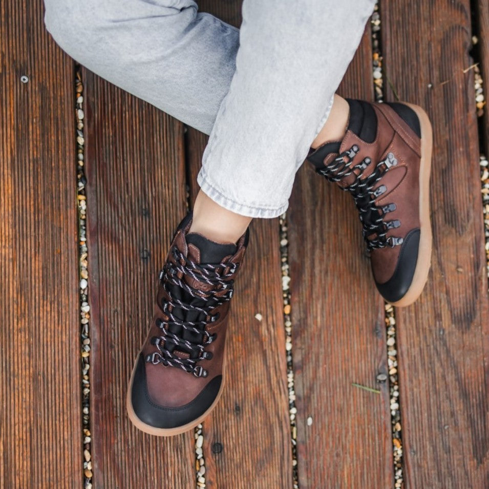 A photo of Be Lenka Ranger boots made with oiled nubuck leather, fleece, and rubber soles. The boots are a hiking boot style and are brown in color with black trim and a fleece lining. A woman is shown from mid leg down sitting with her legs crossed on a porch wearing rolled jeans and the ranger boots.  #color_dark-brown
